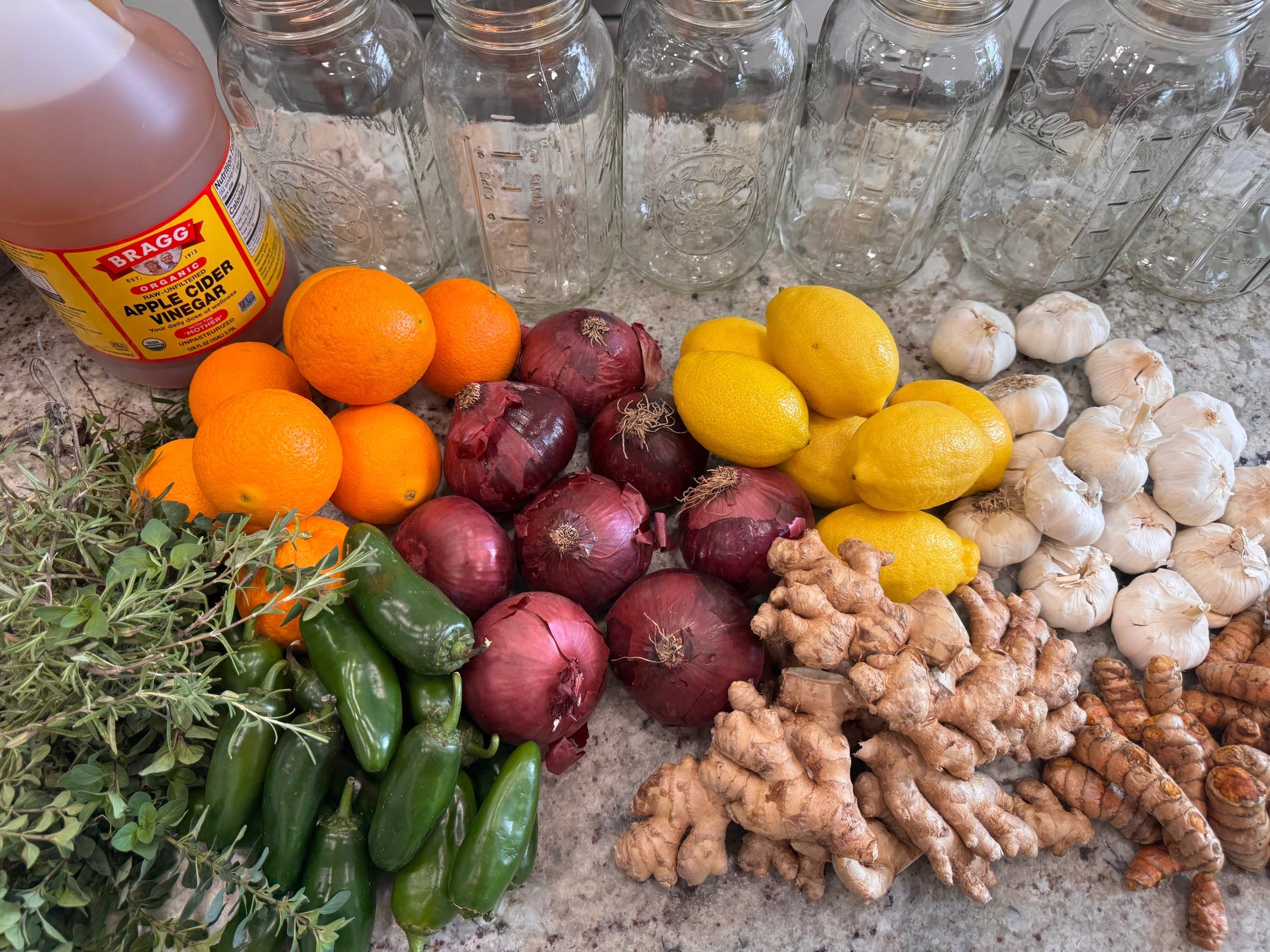 Fire Cider ingredients laid out on a counter: oranges, red onions, lemons, garlic, ginger, turmeric, jalapeños, rosemary, oregano, and apple cider vinegar with empty half-gallon jars